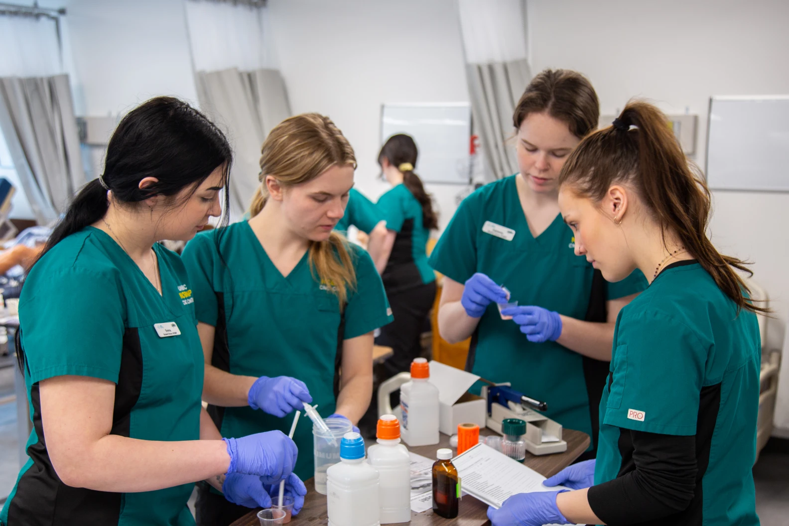 Four nursing students in teal scrubs are standing around a central table and engaged in lab work.